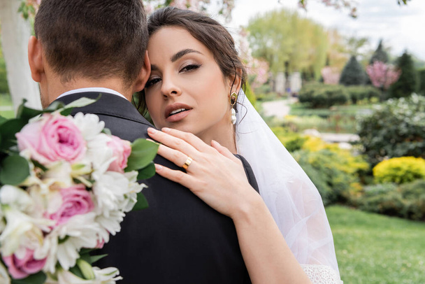 Pretty bride in veil holding flowers and hugging groom in park  - Foto, immagini