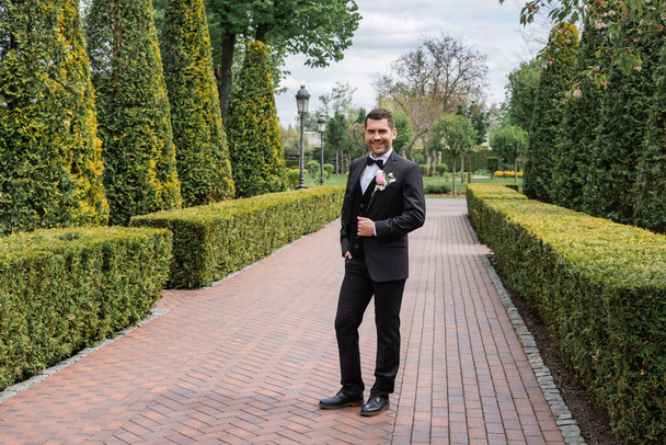 Cheerful groom holding hand in pocket of pants in park  - Foto, immagini