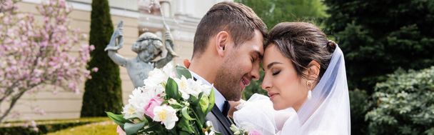 Vue latérale de la mariée avec bouquet embrassant marié souriant près fontaine floue, bannière  - Photo, image