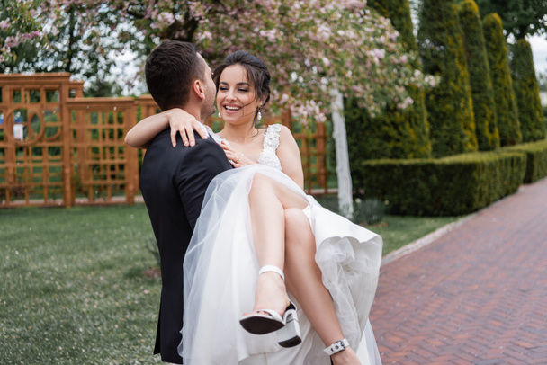 Groom holding on hands smiling bride in white wedding dress in park  - Фото, зображення