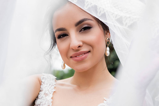 Mosolygó menyasszony néz kamera közelében elmosódott fehér fátyol  - Fotó, kép