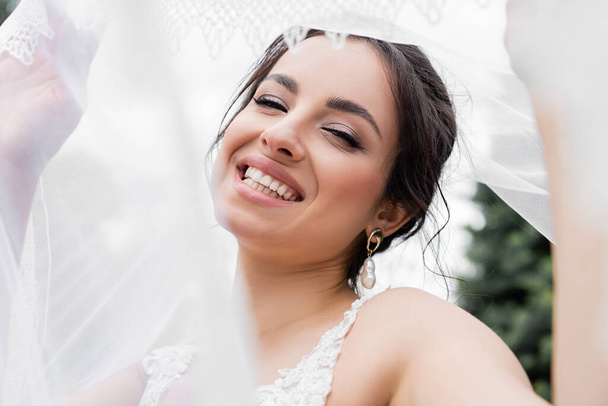 Positive Braut lächelt und hält verschwommenen Schleier im Freien  - Foto, Bild