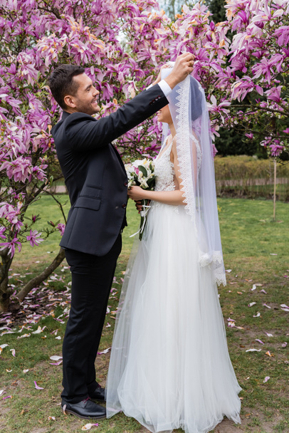 Vue latérale du marié souriant enlevant le voile de la mariée près des magnolias  - Photo, image