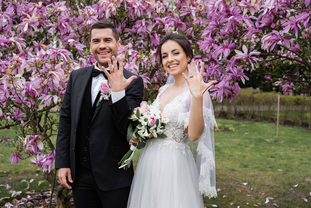 Χαμογελαστή νύφη και γαμπρός που δείχνουν δαχτυλίδια κοντά σε δέντρα μανόλια  - Φωτογραφία, εικόνα
