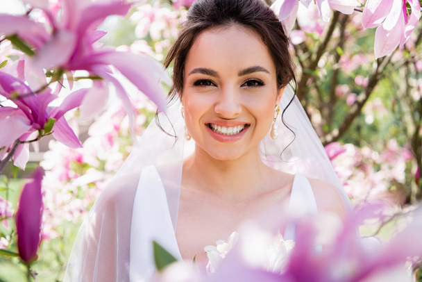 Szczęśliwa panna młoda w welonie uśmiechnięta do kamery w pobliżu kwitnącej magnolii  - Zdjęcie, obraz