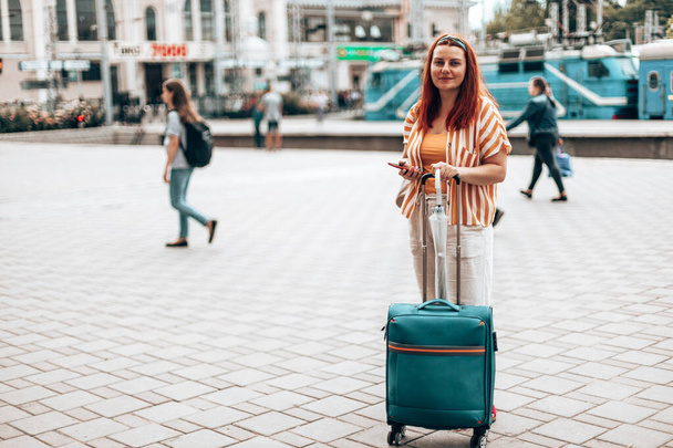 Young caucasian girl in stylish clothes with a smartphone and carry-on luggage stands at the station waiting for the train - Foto, Bild
