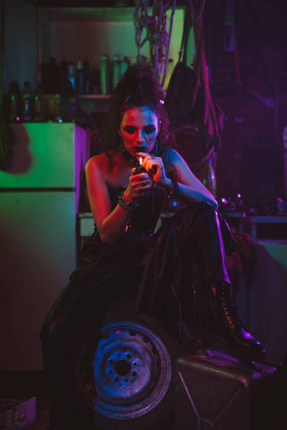 Mädchen im futuristischen Cyberpunk-Kostüm raucht in der Garage eine Zigarette mit Neonlicht. Steampunk-Stil - Foto, Bild