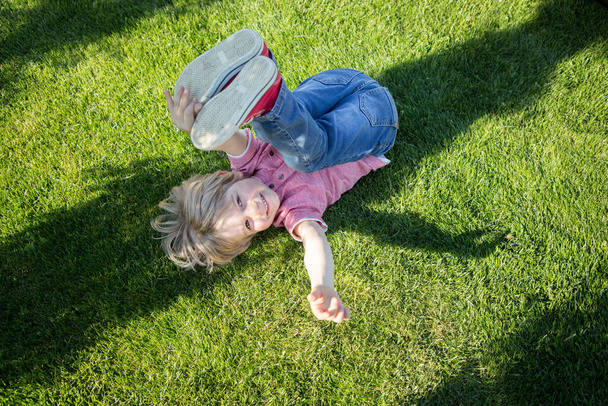 Щасливий маленький хлопчик лежить догори ногами на зеленій траві в парку. Дитяче щастя, радість, самопочуття. Гарний настрій, розслабся
 - Фото, зображення