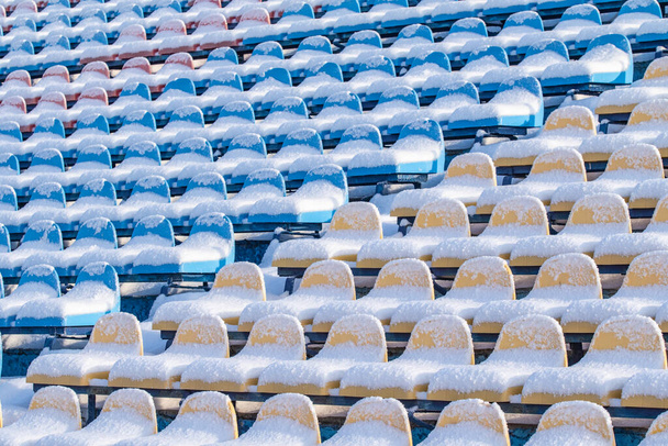 Места на стадионе в снегу зимой. Заснеженные разноцветные сиденья на стадионе зимой. Снегопад мешает спорту. Неочищенный стадион. - Фото, изображение