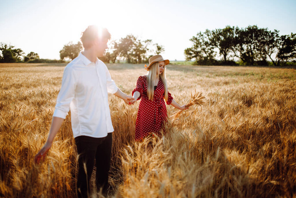 Mladý pár chodí při západu slunce na pšeničném poli. Dívka v hrachových šatech a slaměném klobouku a mladý muž v bílé košili na pozadí modré oblohy. Letní krajina.Dva milenci na pšeničném poli - Fotografie, Obrázek