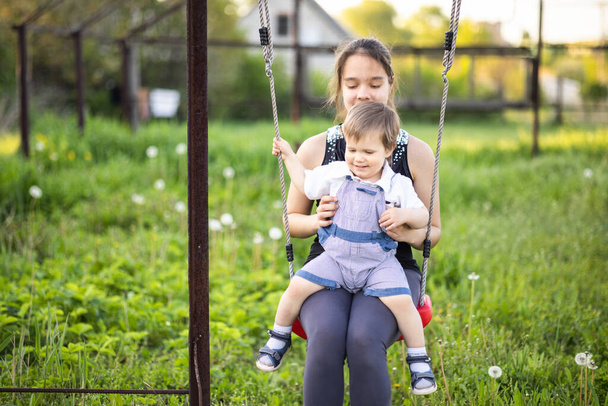 Cute brother and older sister ride on a bright swing on warm spring day against the backdrop of a green blooming garden - Φωτογραφία, εικόνα