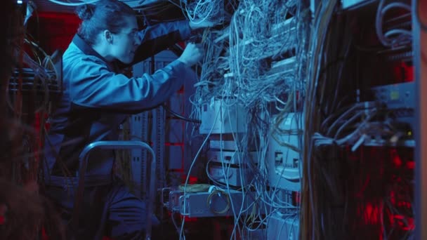 Tilting up of young Caucasian workman wearing glasses, standing on stepladder, repairing hardware in server room, using screwdriver - Footage, Video