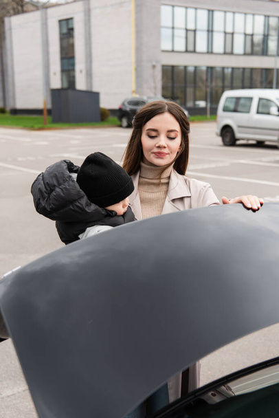 молодая женщина с малышом сыном закрывает багажник автомобиля на открытом воздухе - Фото, изображение