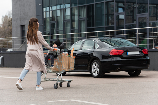 young mother with purchases and toddler son in shopping cart walking on car parking - Photo, Image