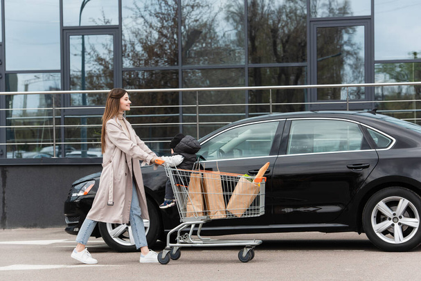 side view of young woman with baby boy and shopping bags in cart on car parking - Foto, Imagem