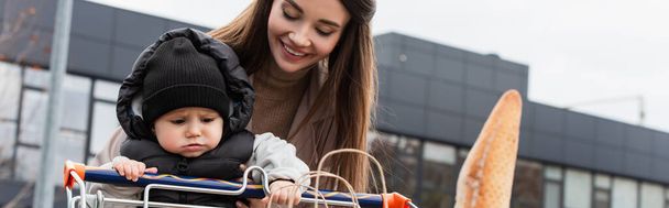 young mother smiling near toddler son sitting in shopping cart, banner - Photo, image