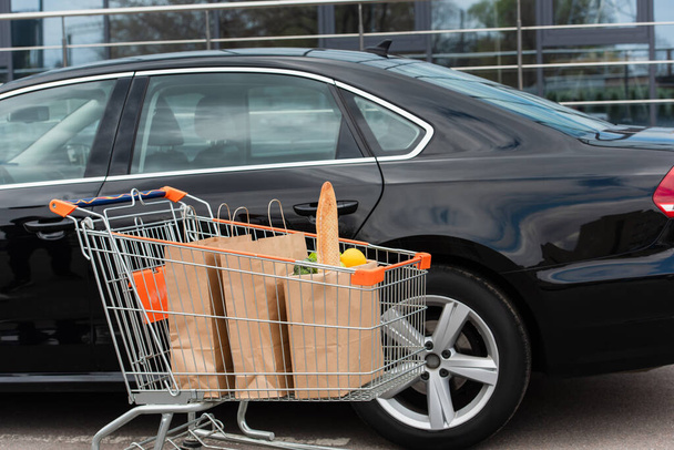 black car near shopping trolley with purchases on parking - Фото, изображение