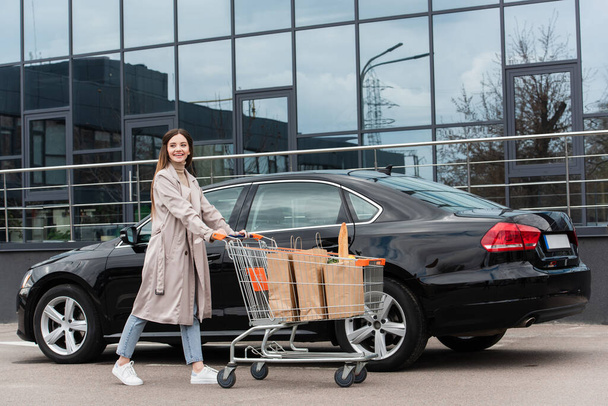 χαρούμενη γυναίκα με τα πόδια τρόλεϊ ψώνια κοντά στο μαύρο αυτοκίνητο στο πάρκινγκ - Φωτογραφία, εικόνα