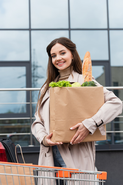 happy woman looking at camera while holding shopping bag with fresh food outdoors - 写真・画像