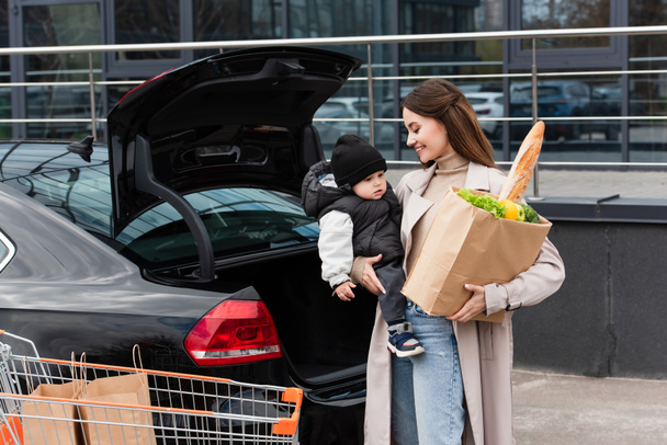 gelukkig vrouw met zoon in handen en vers eten in boodschappentas in de buurt van auto buiten - Foto, afbeelding