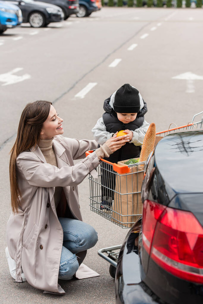 smiling woman giving orange to toddler kid sitting in shopping cart - Fotoğraf, Görsel