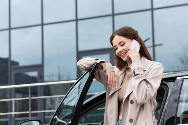young woman smiling while talking on cellphone near automobile - Photo, Image