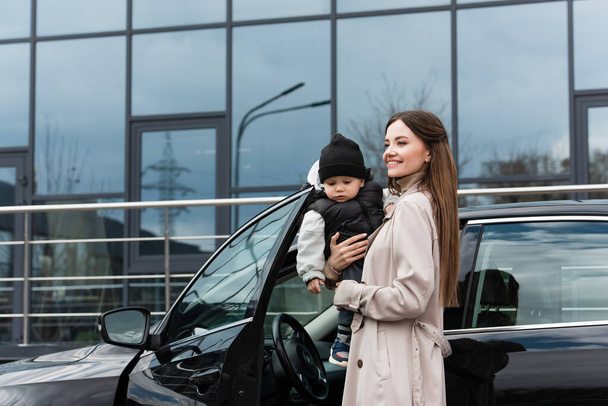 joyful woman looking away while standing with toddler son near car - Foto, afbeelding