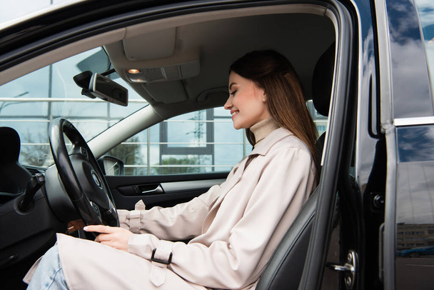 smiling woman in trench coat sitting in car at steering wheel - Foto, Bild