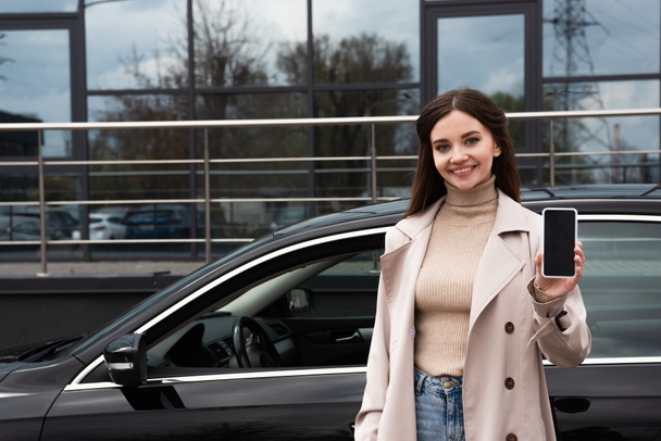radosna kobieta pokazuje smartfon z pustym ekranem w pobliżu samochodu na zewnątrz - Zdjęcie, obraz