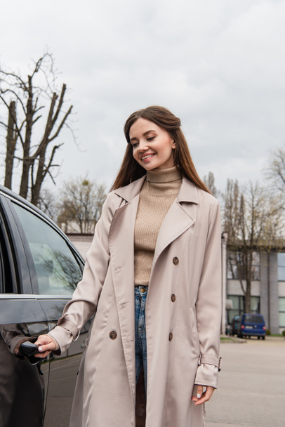 щаслива жінка в траншеї пальто відкриває двері автомобіля
 - Фото, зображення