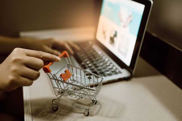 Hand of woman holding trolley shopping cart on table with laptop for shopping. Ideas for shopping online or e-commerce from internet. - Photo, Image