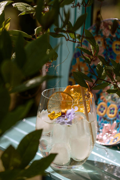 Letni orzeźwienie aperitif koktajl w szkle z lodem, bambusowa słoma i jadalne kwiaty na stole w tropikalnym ogrodzie letnim  - Zdjęcie, obraz