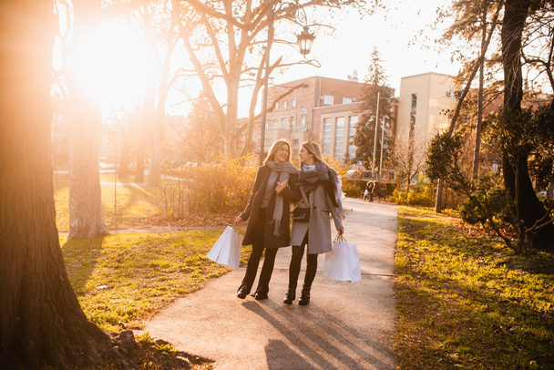 Full length cheerful girls in coats walk through the park with bags in their hands and chat. Blonde sisters in autumn shopping - Fotoğraf, Görsel