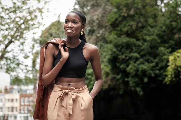 Porträt einer schwarzen Frau in Mode, die mit Lederjacke posiert. Sie steht mitten auf einer Straße und lächelt.. - Foto, Bild