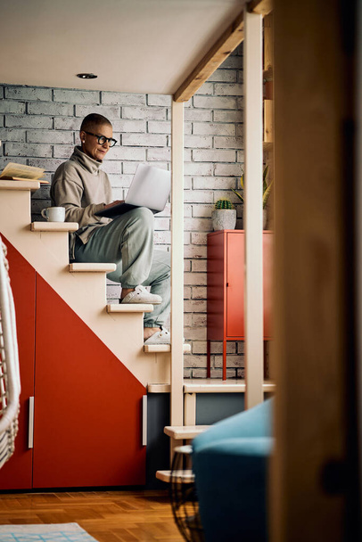 Houkutteleva vanhempi nainen istuu portaissa kotona ja käyttää kannettavaa tietokonetta roikkuu sosiaalisessa mediassa tai työssä. - Valokuva, kuva