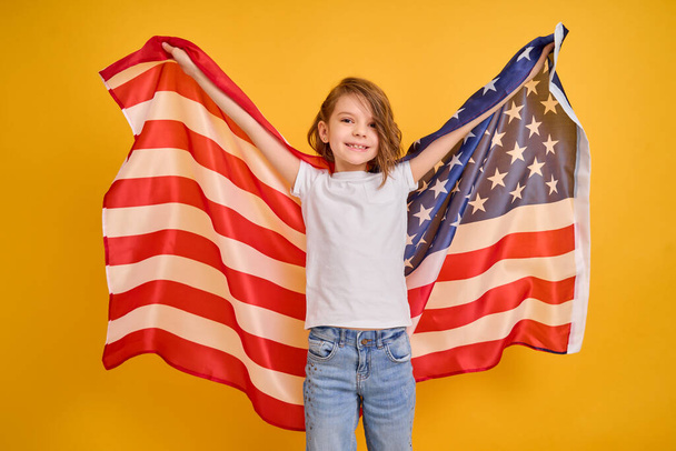 Щаслива дитина, мила дівчина з американським прапором на жовтому тлі студії. США святкують 4 липня День незалежності. - Фото, зображення