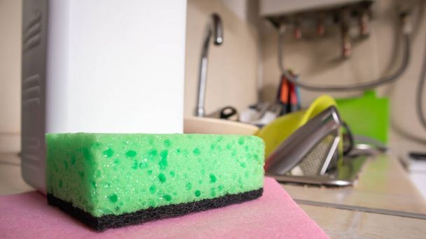 Πράσινο σφουγγάρι και δοχείο υγρού σαπουνιού για το πλύσιμο των πιάτων σε ένα βρώμικο νεροχύτη εντελώς με πιάτα και σκεύη κουζίνας. Πλένει πιάτα στην κουζίνα με το χέρι με απορρυπαντικό και σφουγγάρι - Φωτογραφία, εικόνα