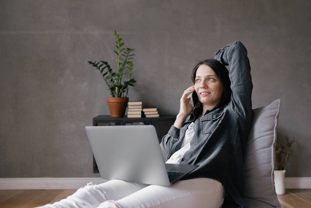top view, European business woman in a gray shirt and white jeans sitting in a chair, working with a laptop and chatting on the phone, texting, internet surfing or doing online shopping - Foto, Imagen