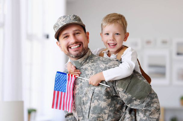 米軍の制服を着た幸せなアメリカ人家族の父親の肖像と、カメラで抱き合って笑っている米国の国旗を持つかわいい男の子の息子、男性兵士の父親は自宅で家族と再会しました - 写真・画像