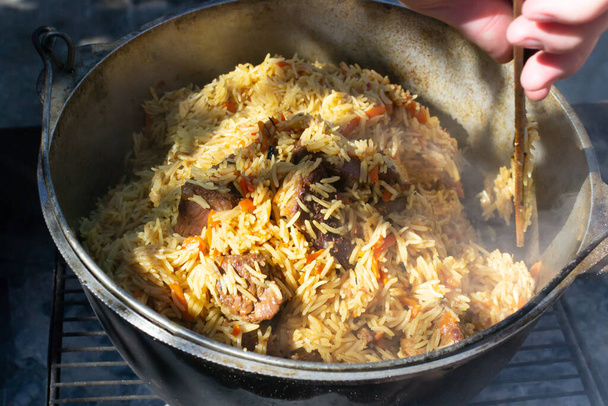 Cooking traditional oriental pilaf over the fire. The man mixes ready-made loose pilaf with vegetables and meat in a cauldron before serving. - Foto, Imagen