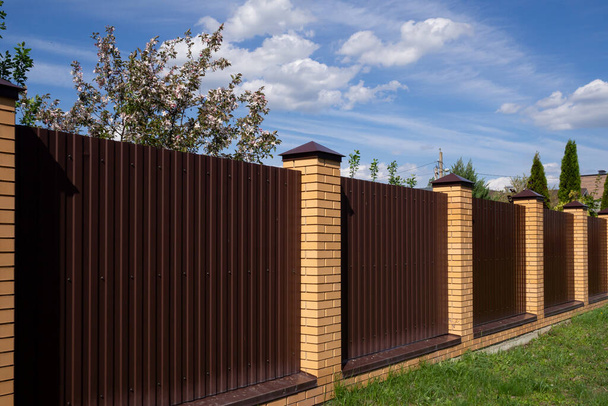 A brown metal fence with brick posts against a blue sky. A high wall encloses the private area. Security concept. Copy space - Zdjęcie, obraz