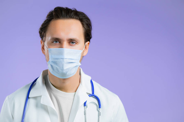 Portrait of doctor in professional medical coat and facial mask. Man doc isolated on purple background. Medical staff concept. - 写真・画像