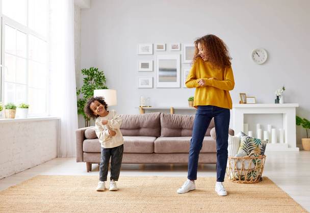 Happy afro american family mother and son dancing with music and playing together while standing in living room at home, full length shot. Mom and child in casual wear enjoying leisure time together - Zdjęcie, obraz