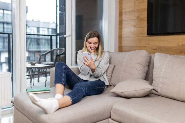 Young beautiful woman sitting at home on the couch and uses a mobile phone, resting from work - Foto, Imagen