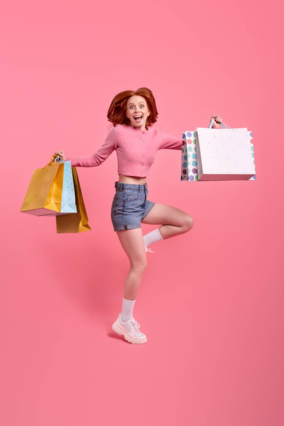 Full-length portrait of redhead female with packages jumping high up celebrating weekend - Фото, зображення