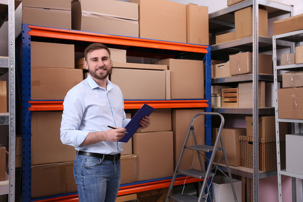 Young businessman with clipboard near rack of cardboard boxes at warehouse - Фото, изображение