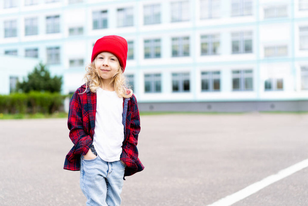 portré egy gyönyörű szőke kislányról az utcán, piros kalapban és egy ketrecben lévő ingben.. - Fotó, kép