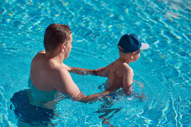 Καυκάσιος πατέρας και γιος στην πισίνα. Ο πατέρας μαθαίνει στο γιο του να κολυμπάει στην πισίνα μια ηλιόλουστη μέρα. Να κάνουμε παρέα. Καλοκαίρι διασκέδαση - Φωτογραφία, εικόνα