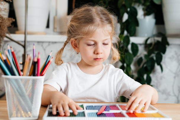 家のテーブルでモデルをするための色のプラスチックの明るいカラフルなパレットを持つ小さなブロンドの女の子、趣味と子供の発達. - 写真・画像