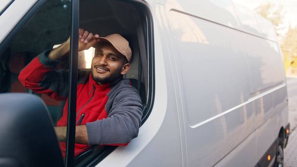 Ένας ντελιβεράς με κόκκινη στολή που κάθεται στο άσπρο φορτηγάκι και δίνει φιλοδώρημα στο καπέλο του. - Φωτογραφία, εικόνα